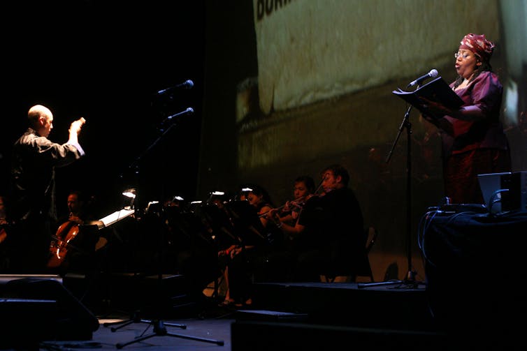 Wide shot. A woman dressed in purple, her hair covered by a traditional cloth and wearing glasses, sings into a mic as she reads music that she holds. Opposite her is a conductor in a spotlight and between them an ensemble of classical musicians.