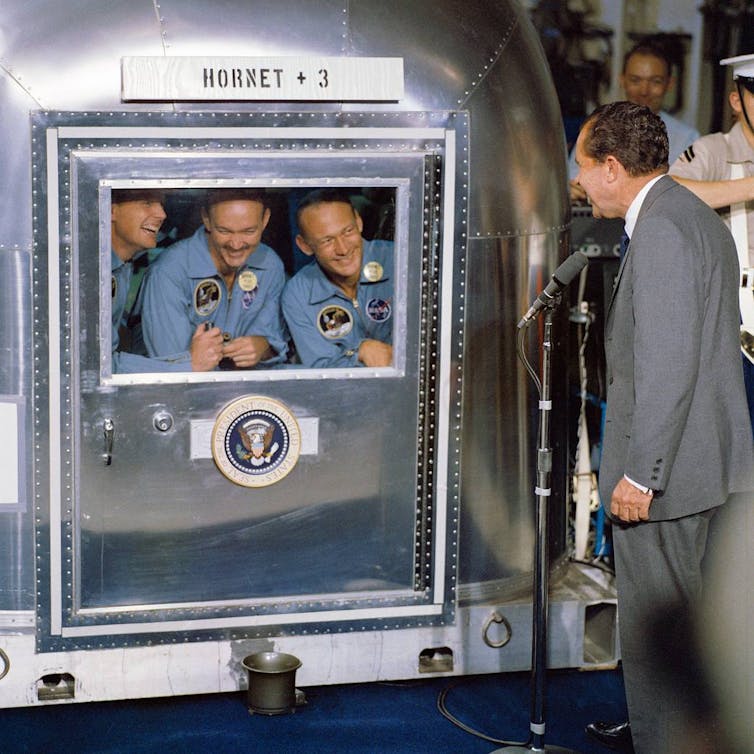 Apollo 11 astronauts in a small metal quarantine facility, with President Richard M. Nixon speaking to them from outside.
