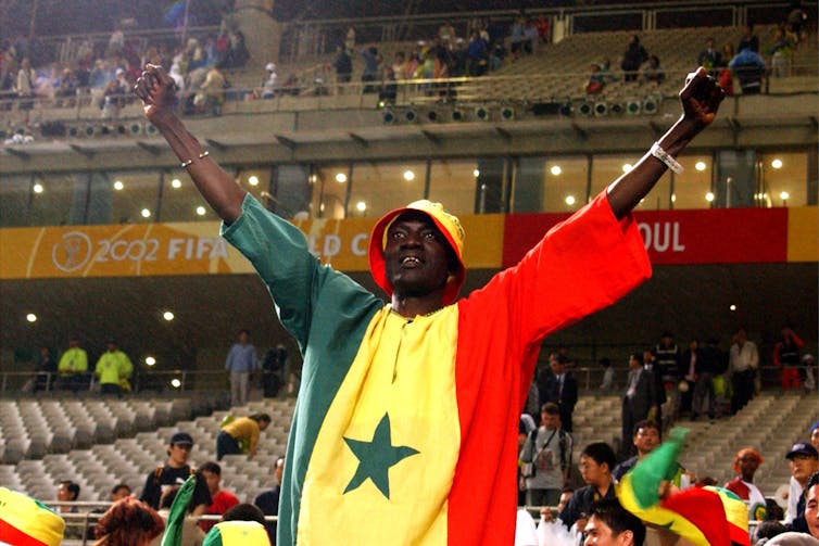 A man stands with two fists in the air, wearing a shirt comprised of the colours of the flag of Senegal - green, yellow and red, behind him a sports stadium.