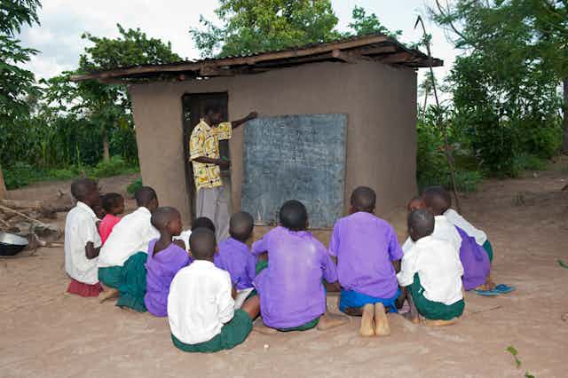 Children sit on the ground outside a small building while a man points to a blackboard on the exterior wall of the building