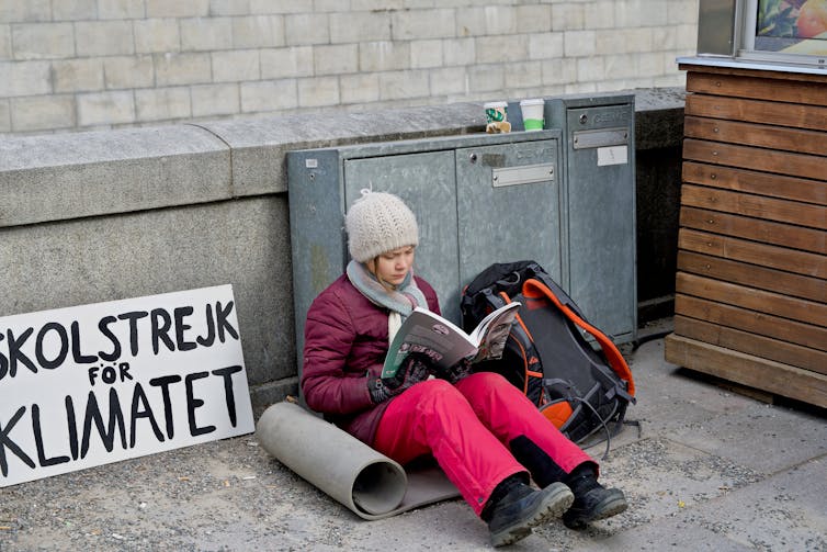 Greta Thunberg sits reading with her famous sign nearby.