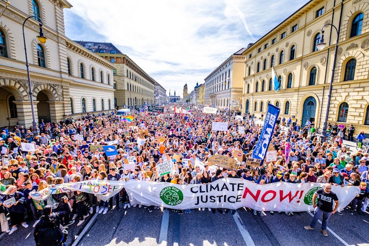 A vast crowd holding protest banners march along a wide city street.