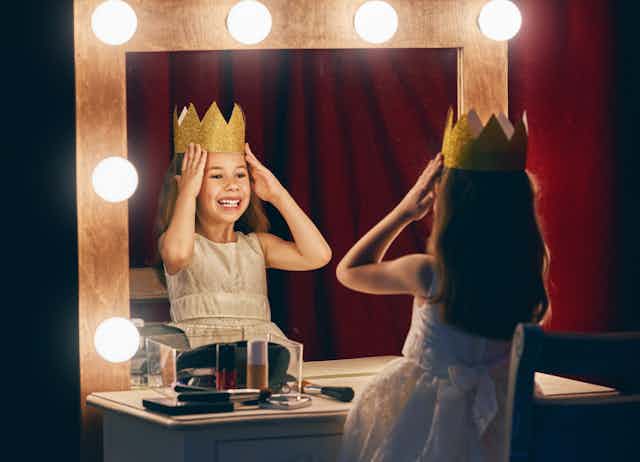 A young girl places a crown on her head while looking in the mirror. 