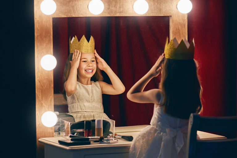A young girl places a crown on her head while looking in the mirror.