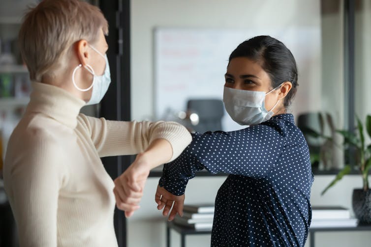 Two women in masks greeting one another by bumping elbows