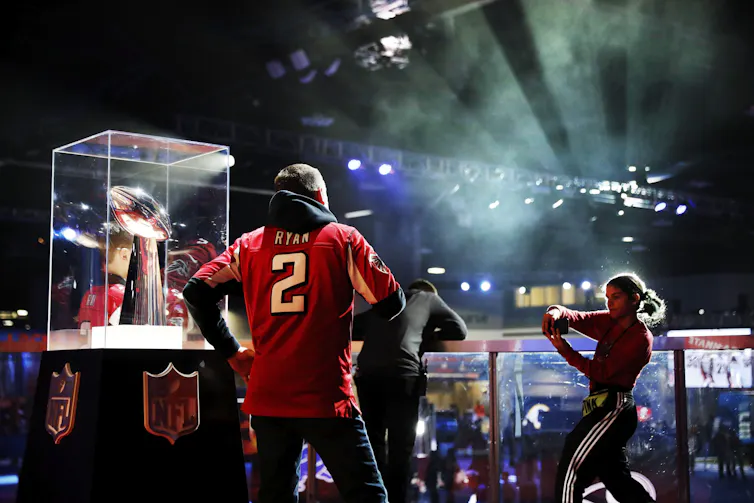 A man standing next to a trophy in a glass case, while a women takes a photo of him with her phone.