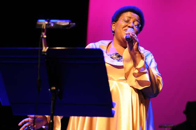 Against a pink backdrop and in a gold dress, a woman with a short Afro and glasses sings into a microphone, her eyes closed.
