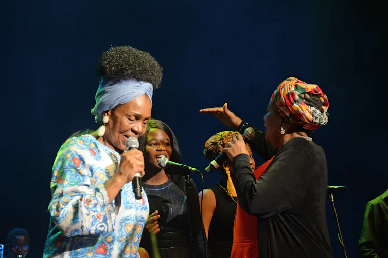 Four women on stage, two backing singers - who a woman instructs using a hand gesture and an elder woman foregrounded, singing into a microphone.