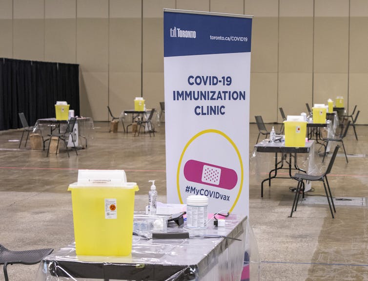 A large empty room with tables set up with vaccination supplies and biohazard bins.