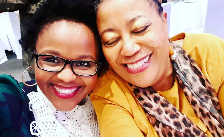 Two women smiling into a camera, one in glasses and the other, eyes closed in a mustard blouse with leopard print scarf, looking happy and bonded.