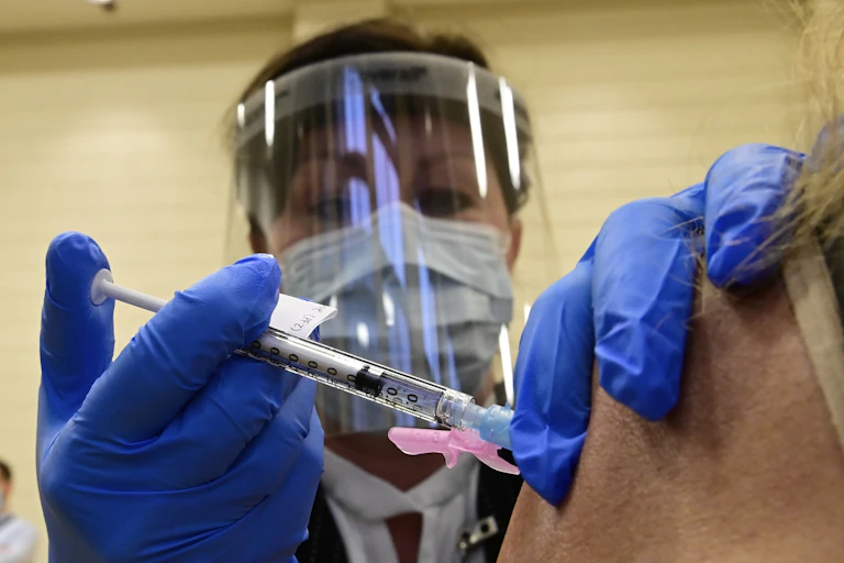 A woman's face in a shield and mask, with a syringe injecting someone's shoulder in the foreground.