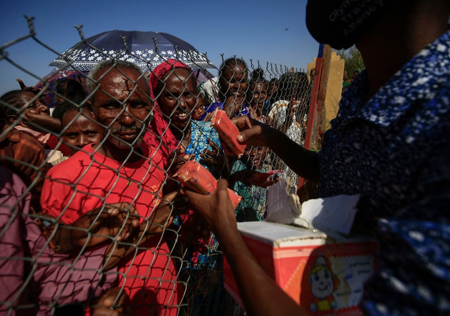 A group of people behind a wire fence reach for food items being handed over by a person in front of the fence