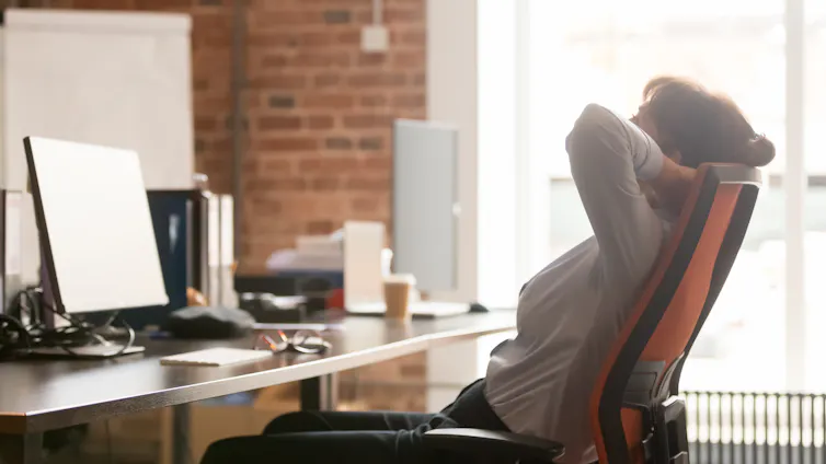 An office worker in a chair sits back with their hands hend their head looking at their screen