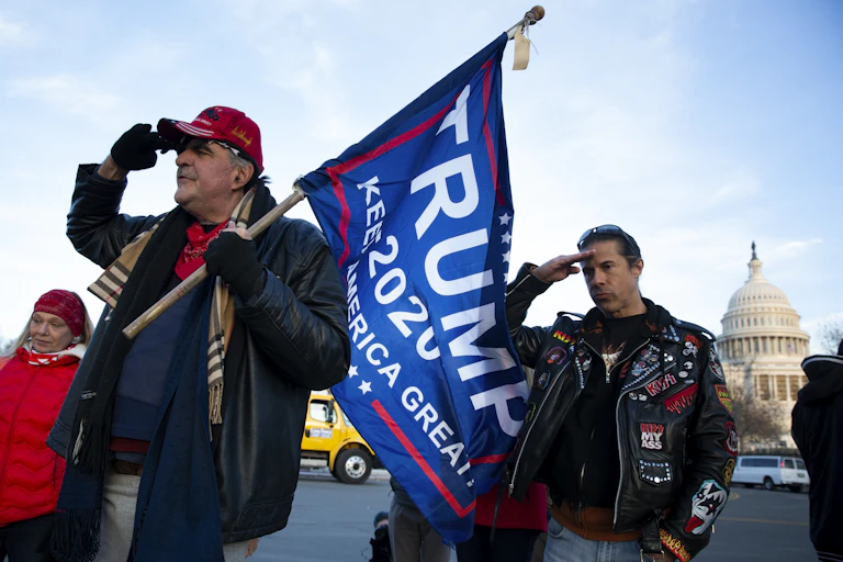 Two men and a woman, one carrying a Trump 2020 flag saluting with the US Capitol in the background