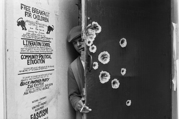 An unidentified member of the Black Panthers peeks around a bullet-pocked door that police blasted with gunfire during a predawn raid.