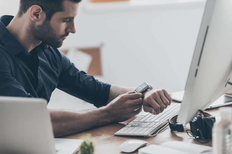 A man sat in front of a desktop computer checks his smart watch