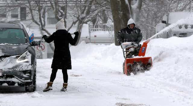 A woman scrapes snow off her car while a man uses a snowblower to clear his driveway. 