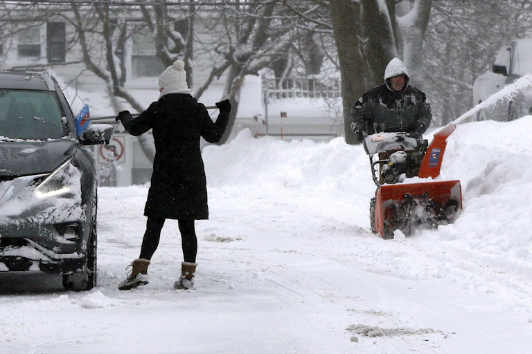 A woman scrapes snow off her car while a man uses a snowblower to clear his driveway.