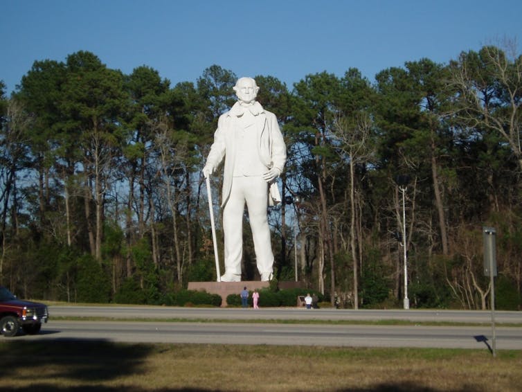 A giant white marble statue of a man with a cane, highway in the foreground