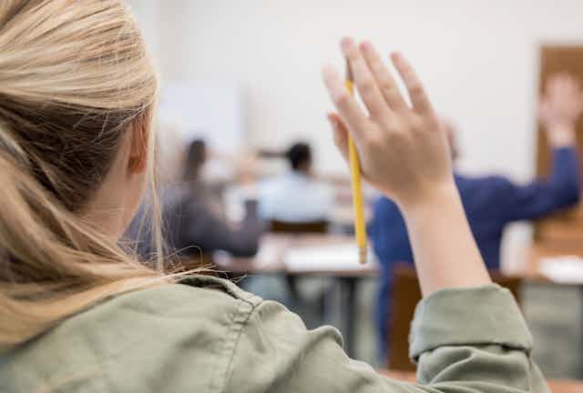 A girl holds up her hand, which is holding a pencil, in class 