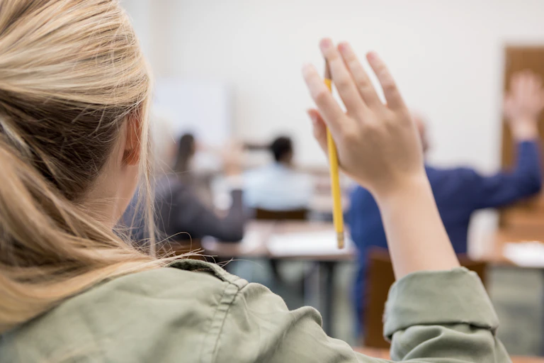 A girl holds up her hand, which is holding a pencil, in class