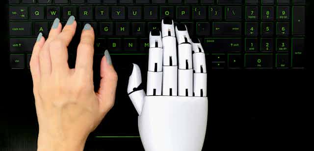 Robot hand typing next to human hand on computer keyboard.