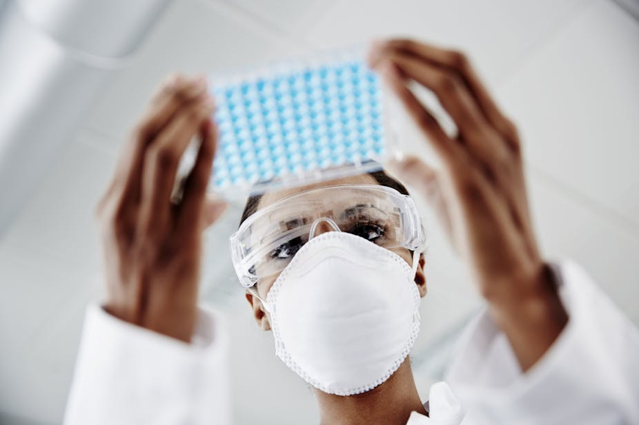 A woman in medical goggles and a white face mask is holding a rectangular dish holding laboratory samples.