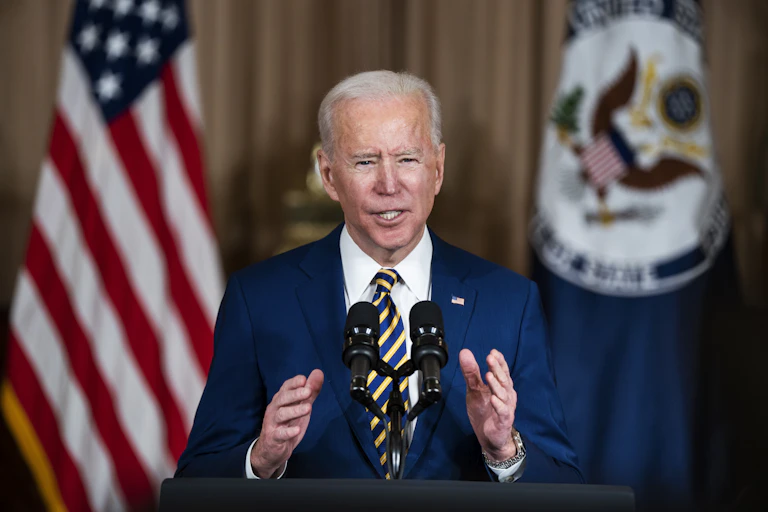 A grey-haired man wearing a blue suit speaks a podium, with a US flag in the background