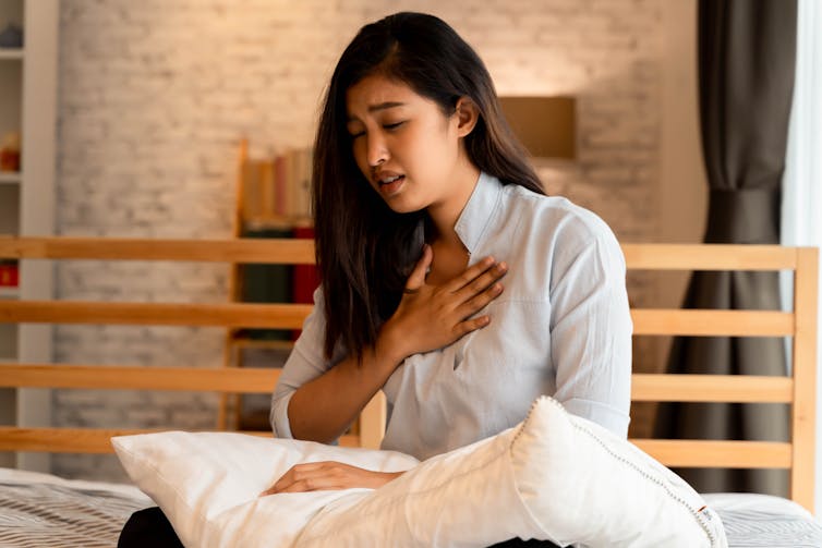 A woman sits on bed holding a pillow and touching her chest.