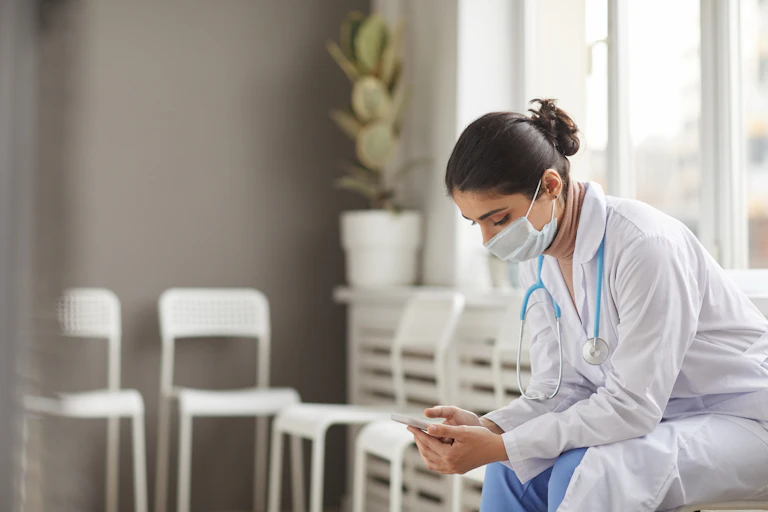 Nurse in mask and in white coat using her mobile phone during her break sitting on chair at the corridor
