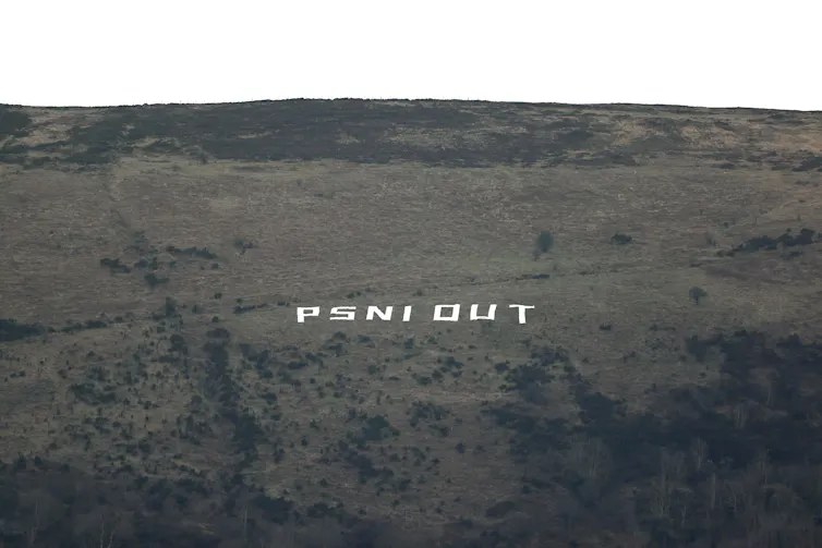White lettering laid out on the side of a hill in the open air spells out 'PSNI Out', referring to the Police Service of Northern Ireland.