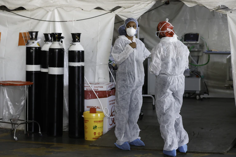 Two healthcare workers in full PPE stand next to oxygen tanks.