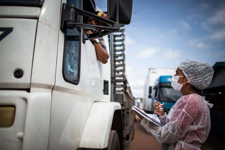 A woman in full PPE interviews a man in the cab of a lorry.