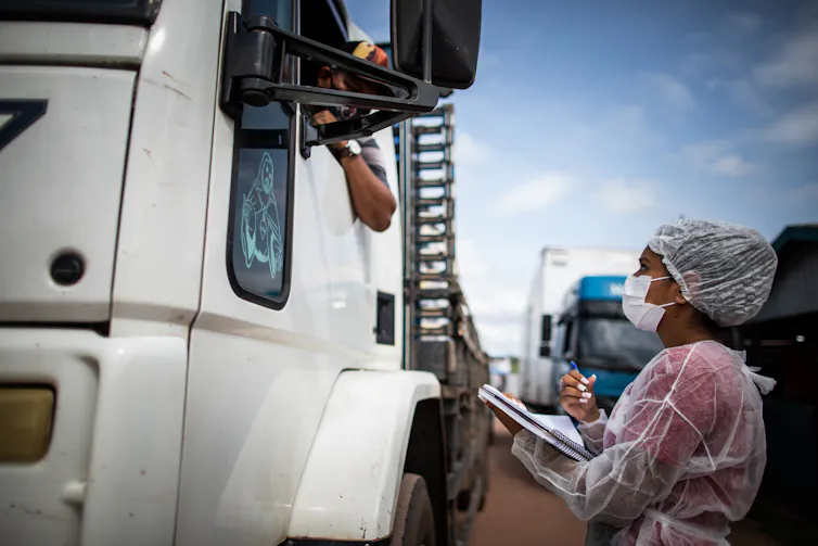 A woman in full PPE interviews a man in the cab of a lorry.