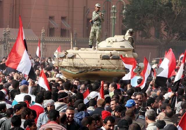 An Egyptian soldier stands on a tank in a square full of protesters waving the Egyptian flag.