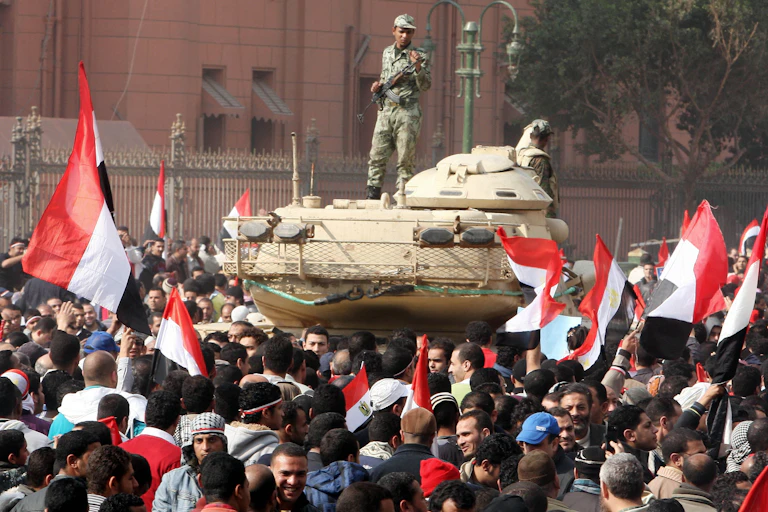 An Egyptian soldier stands on a tank in a square full of protesters waving the Egyptian flag.
