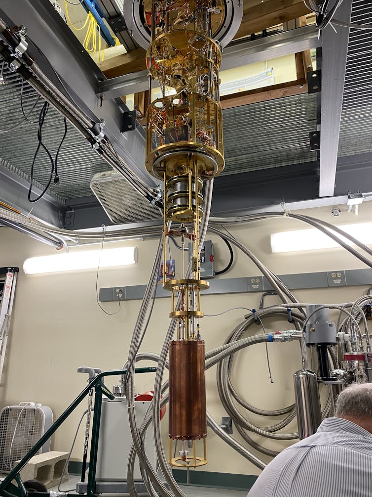 The HAYSTAC detector, a large copper cylinder connected to a gold-plated assembly of tubes and wires hanging from the ceiling of a lab.