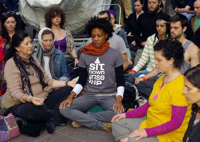People sit on the ground and meditate. One t-shirt reads, "I sit down, I rise up.'