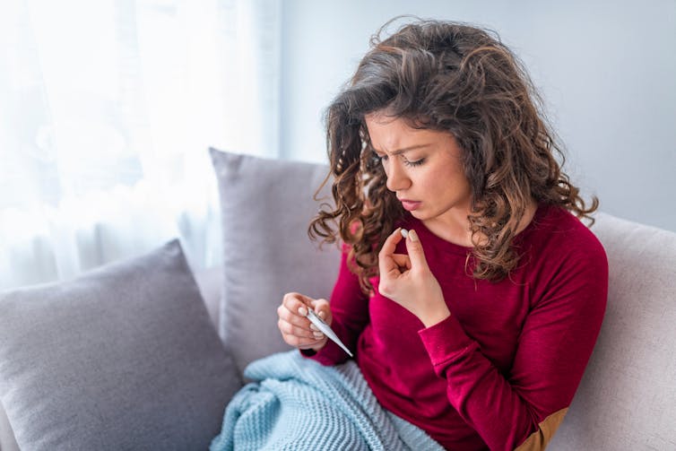 A woman sits on the couch looking at a thermometer and about to take a tablet.