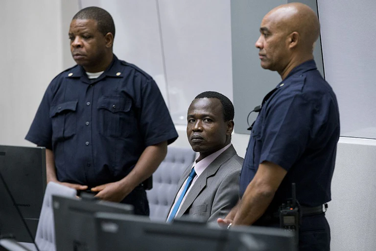 A man in a suit sits between two uniformed standing men