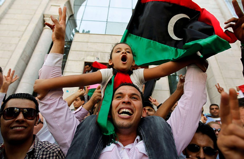 Smiling man with young girl on his shoulders with a Libyan flag in the background.