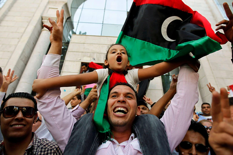 Smiling man with young girl on his shoulders with a Libyan flag in the background.