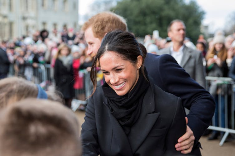 Meghan Markle greeting a crowd with her husband Prince Harry behind her.