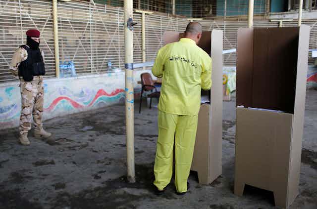 A man wearing a prison uniform casts a vote at a cardboard ballot booth.
