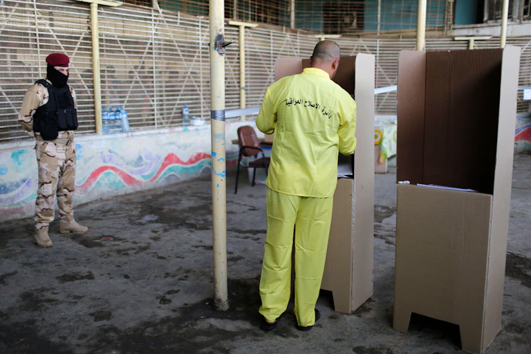A man wearing a prison uniform casts a vote at a cardboard ballot booth.