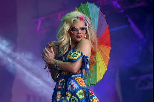 Drag queen on stage wearing a multi-coloured nitted dress and holding a rainbow umbrella