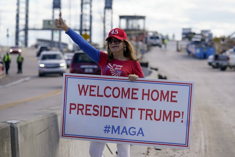 A woman holds a Welcome Home President Trump sign on a roadside.