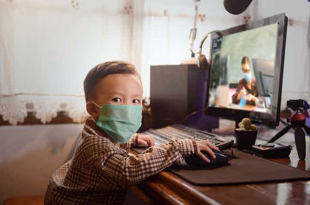 Child wearing face mask sits at computer