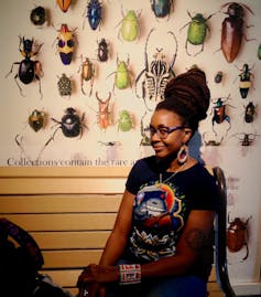 A woman with a wry smile and large hairstyle sits in front of a museum display of insects.