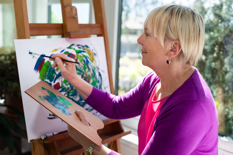 Older woman painting a picture on an easel.